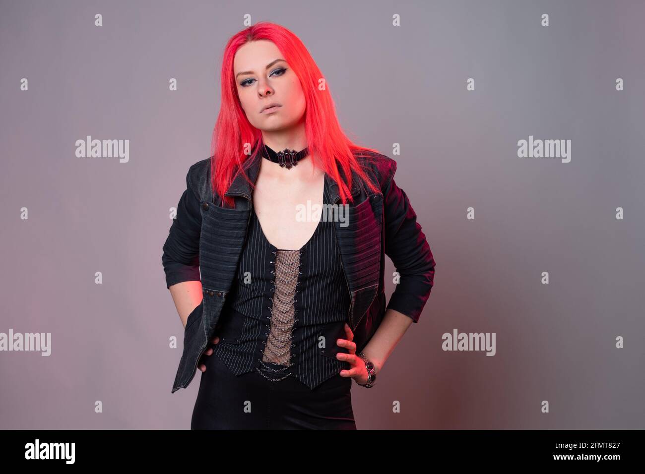 Style kitsch, mode du futur. Mélange de styles. Une jeune femme avec des cheveux écarlate lumineux dans des vêtements sombres inhabituels sur fond gris. Banque D'Images