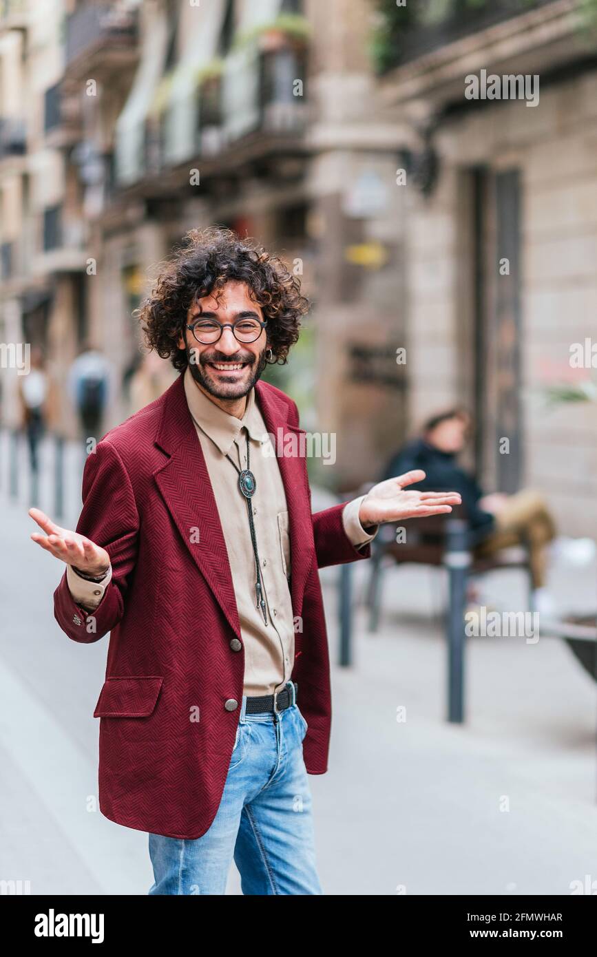 jeune et attrayant homme aux bras ouverts et souriant et regardant la caméra dans la rue. Porte des vêtements et des lunettes tendance. Concept de plaisir, de doute, de questionnement Banque D'Images