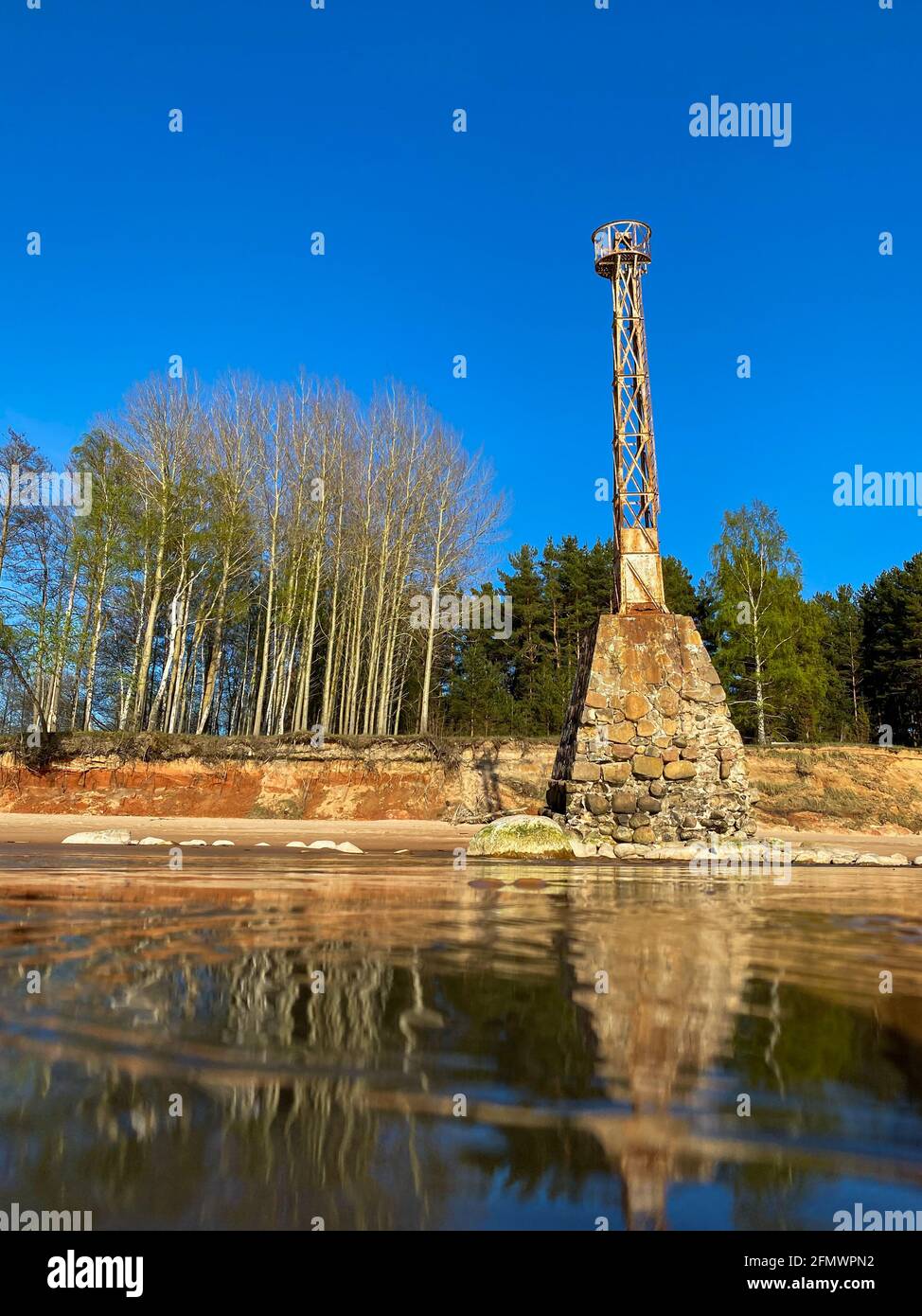 vieux phare en métal rouillé abandonné avec une grande fondation en pierre au bord de la mer avec un reflet dans l'eau. rive de falaises de grès. Pierres de rivage. Banque D'Images