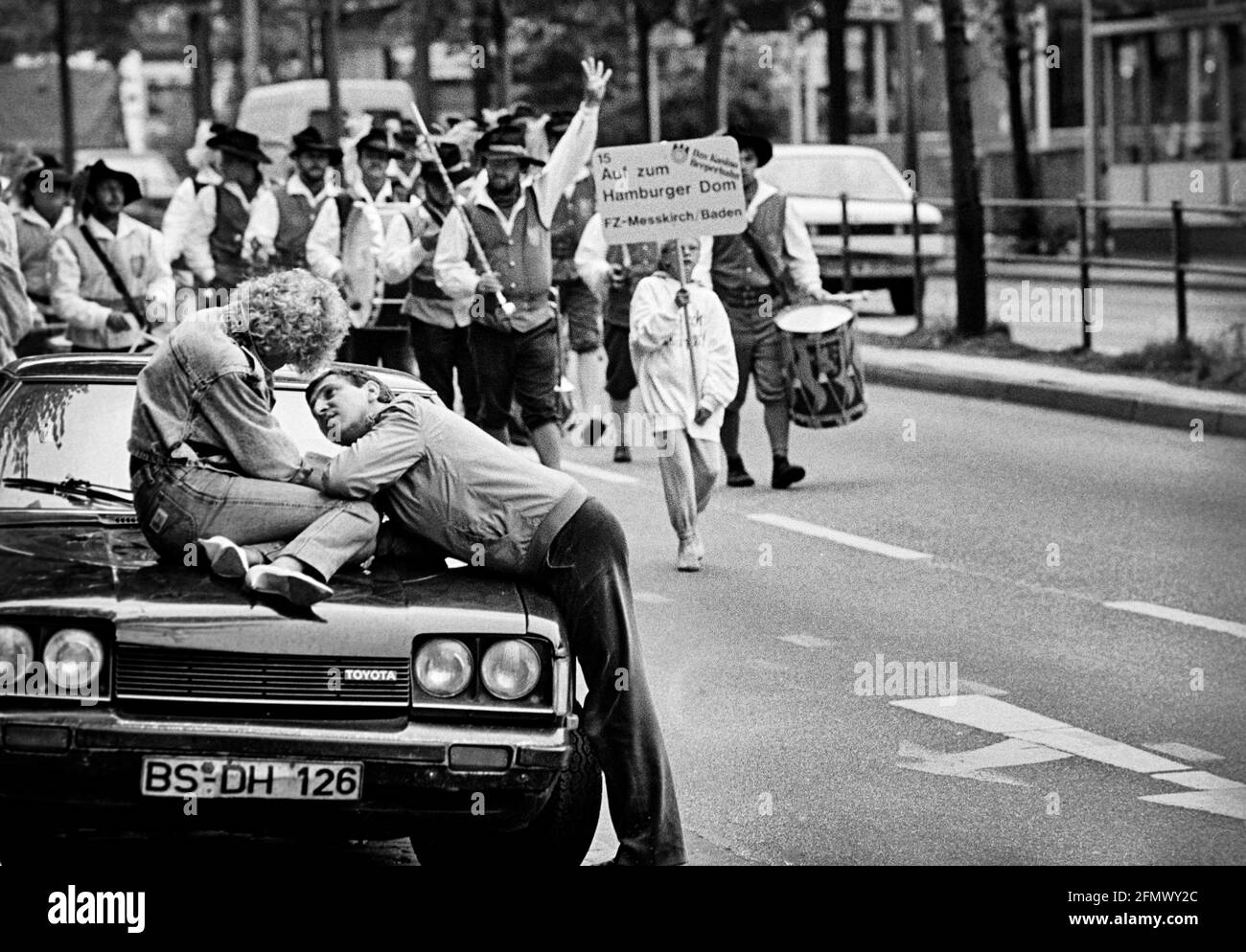Un homme prend soin de la figure incrustées sur le capot de sa voiture, tandis qu'un groupe de marche défichier le Reeperbahn de Hambourg en arrière-plan. 08.08.198 Banque D'Images