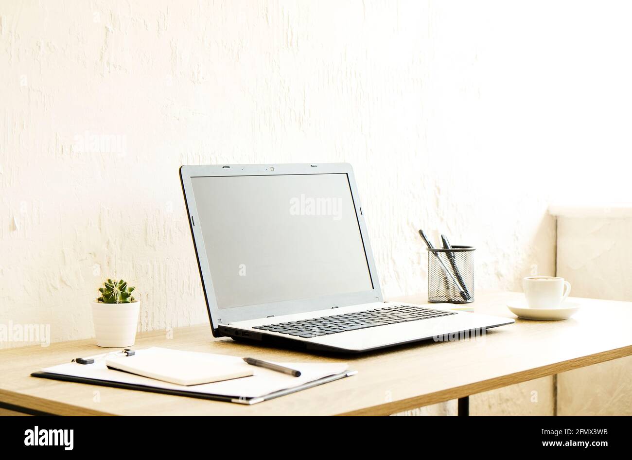 Ordinateur portable à écran vierge, tasse de cappuccino, cactus, fournitures et lunettes pliées sur bureau en bois dans un bureau spacieux plein de lumière du soleil. CR Banque D'Images