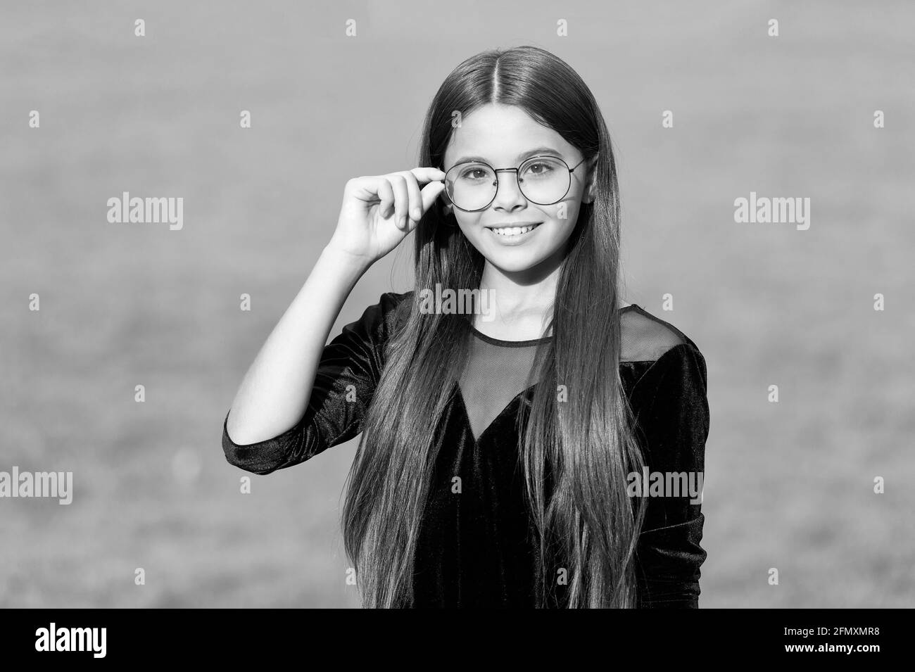 Bonne petite fille avec de longs cheveux dans la mode robe noire fixer verres correcteurs avec beauté look vert herbe ensoleillé été plein air, opticiens Banque D'Images