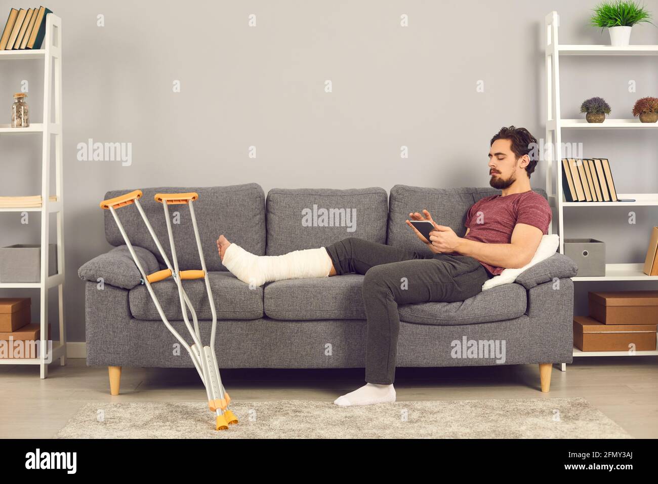 Homme avec une jambe cassée couché sur un canapé et la navigation sur les réseaux sociaux sur une tablette numérique. Banque D'Images
