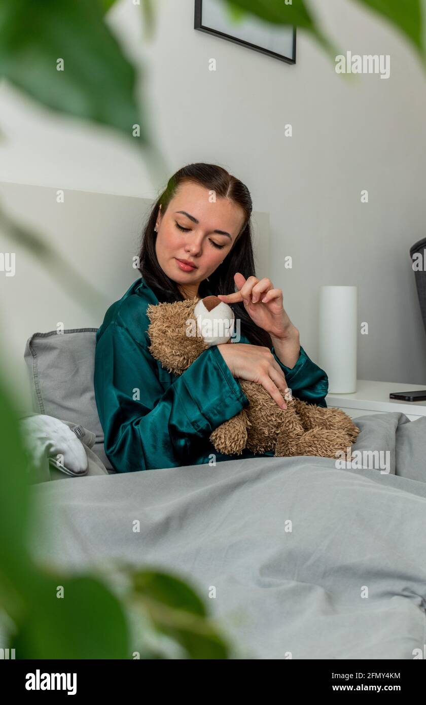 Une jeune femme en pyjama de soie est assise sur le lit et épouse un ours en peluche. Retour aux racines. Concept bien-être. Banque D'Images
