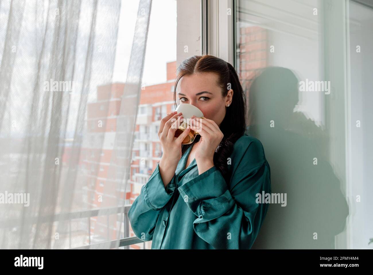Une jeune femme boit du café dans une tasse tout en se tenant près de la fenêtre. Concept de bien-être et zone de confort à la maison. Banque D'Images