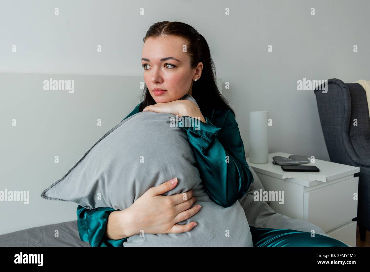 Une jeune femme en pyjama de soie est assise sur le lit et épouse un oreiller. Solitude à l'intérieur de la maison. Concept bien-être. Banque D'Images