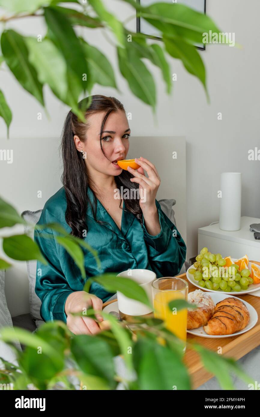Jeune femme en pyjama de soie a un petit déjeuner sain tout en étant allongé sur le lit dans la chambre. Concept de bien-être à la maison. Santé émotionnelle d'une jeune femme Banque D'Images
