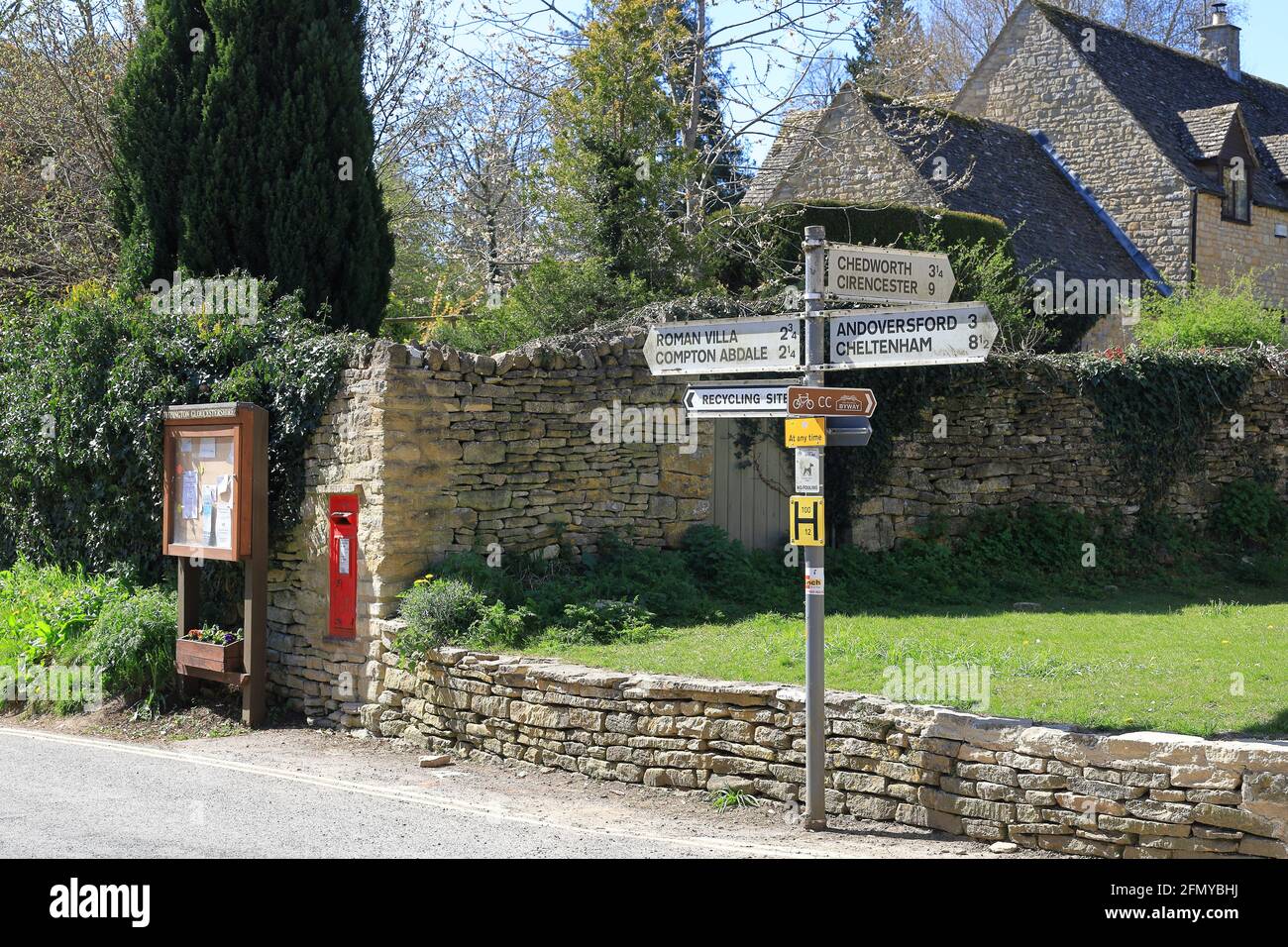 Panneau indiquant la direction avec panneau d'affichage du village et petite boîte postale rouge Royal Mail fixée dans un mur en pierre dans le village de Cotswold de Withington au Royaume-Uni Banque D'Images