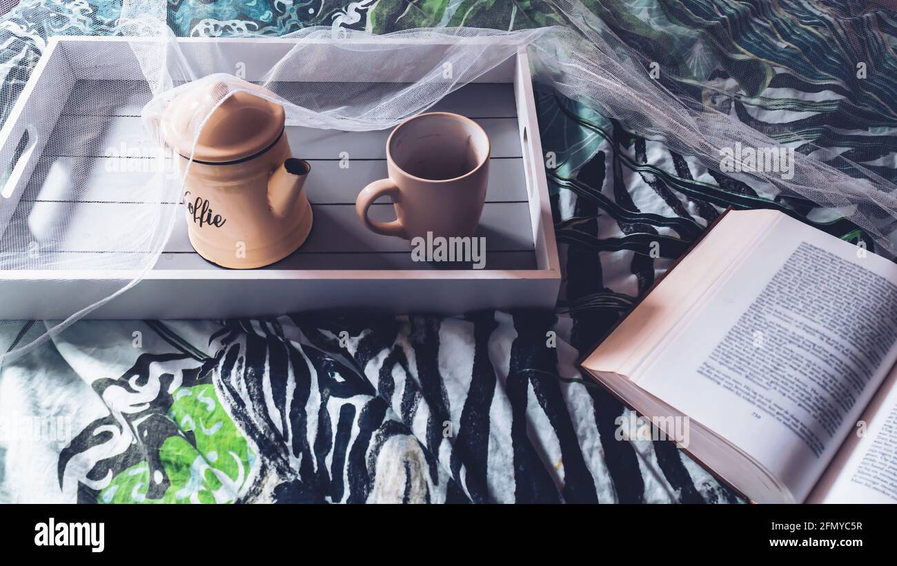 Image d'un plateau en bois avec une cafetière et une tasse assises sur un lit à l'intérieur d'une moustiquaire. Sur le lit est un livre ouvert dans la lumière du matin. Banque D'Images