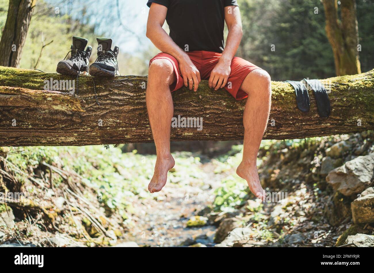 Homme assis sur l'arbre déchu en bois au-dessus du ruisseau de la forêt de montagne pendant qu'il attend des chaussettes séchage de blanchisserie et des bottes de trekking. Personnes actives traveri Banque D'Images