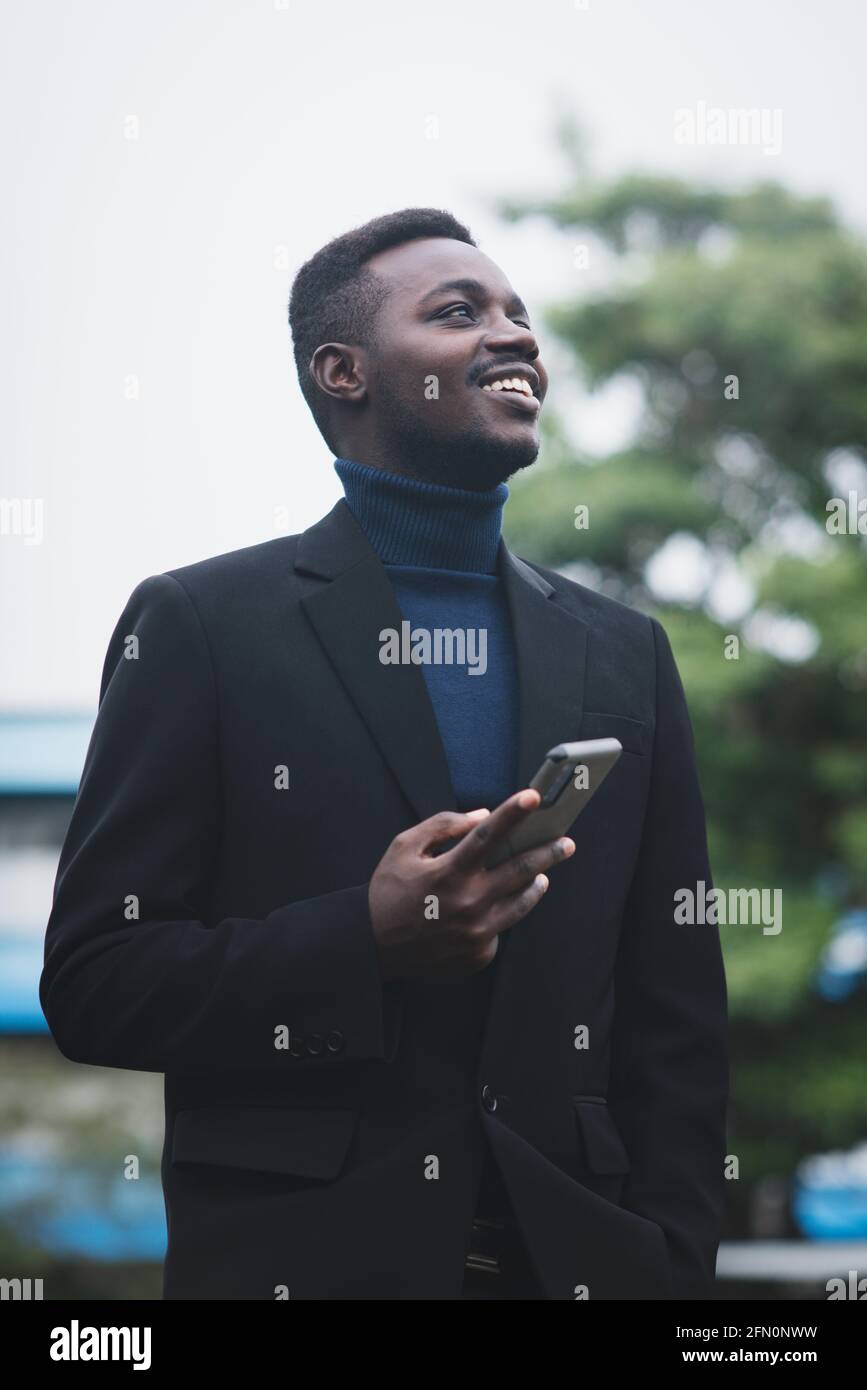 Homme d affaires africain utilisant un smartphone dans un costume noir a la mode. Un homme avec une barbe portant une manche longue ou un pull bleu Photo Stock Alamy