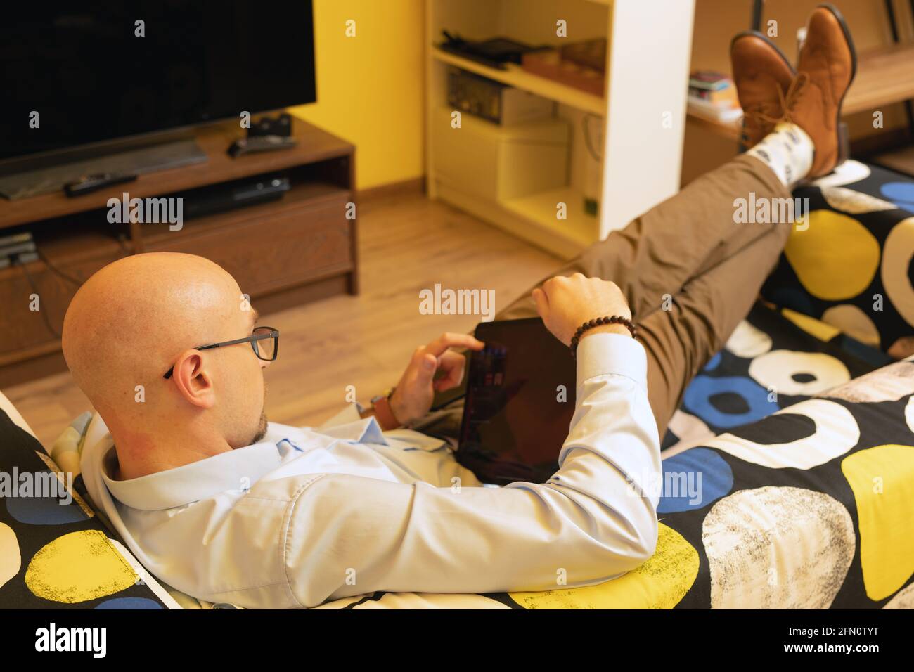 Homme d'affaires chauve avec des lunettes lit le texte ou e-livre tout en étant allongé sur le canapé dans son temps libre. Activités à temps libre. Banque D'Images
