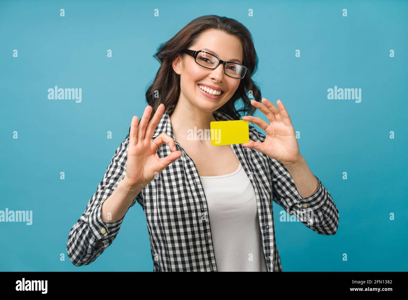 Jolie femme souriante dans une chemise habillée et des lunettes, montrant la carte de crédit à la main pour le concept de société financière et sans cashless, posant sur fond bleu Banque D'Images