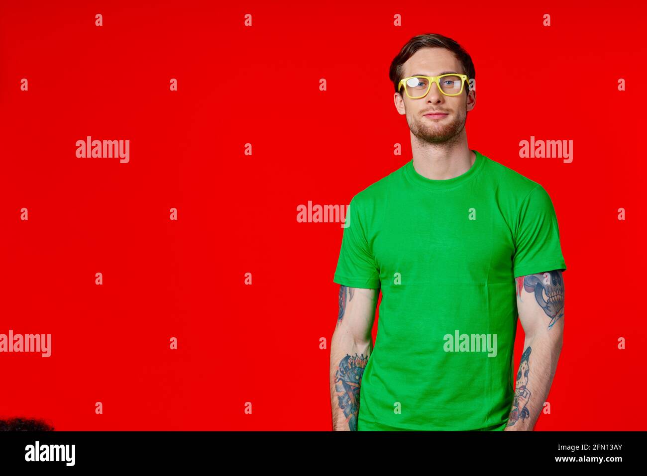 un homme dans un t-shirt vert avec des tatouages dans son mains portant des lunettes sur fond rouge Banque D'Images