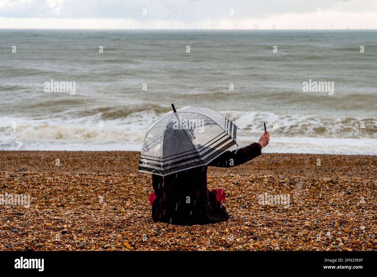 Une jeune femme assise dans la pluie sur Brighton Beach en train de prendre UN selfie, Brighton, East Sussex, Royaume-Uni. Banque D'Images