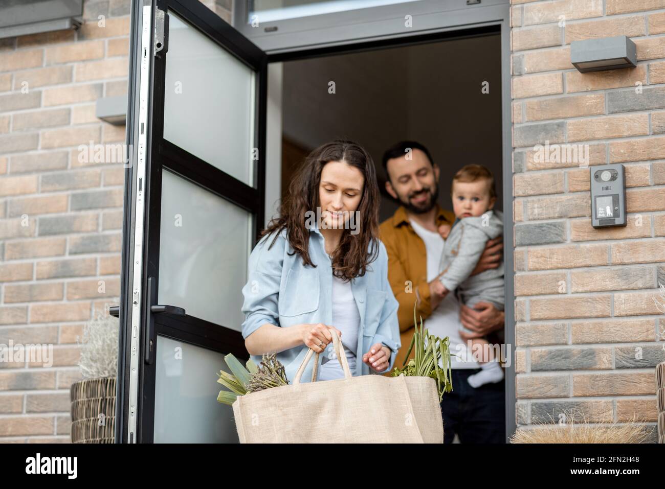 Les jeunes parents avec un nouveau-né enfant client commandent la livraison de nourriture en ligne et debout devant la maison pour la recevoir. La femme au foyer apporte son sac de shopping pendant que son mari tient un bébé. Banque D'Images