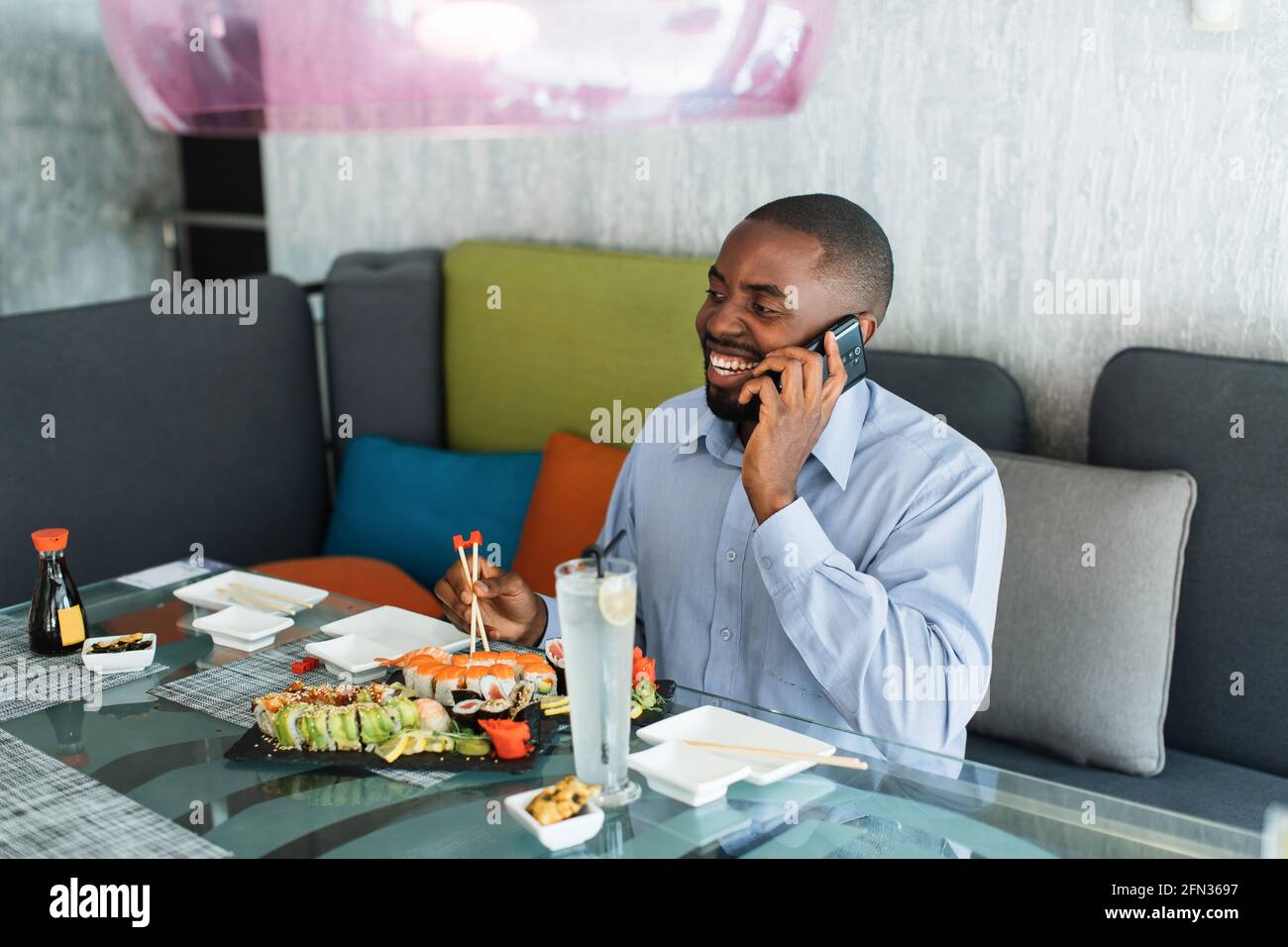 Un Africain est assis dans un restaurant, parlant au téléphone et mangeant des sushis japonais. Un homme d'affaires souriant mange des sushis avec des baguettes et parle avec un ami pendant le déjeuner Banque D'Images