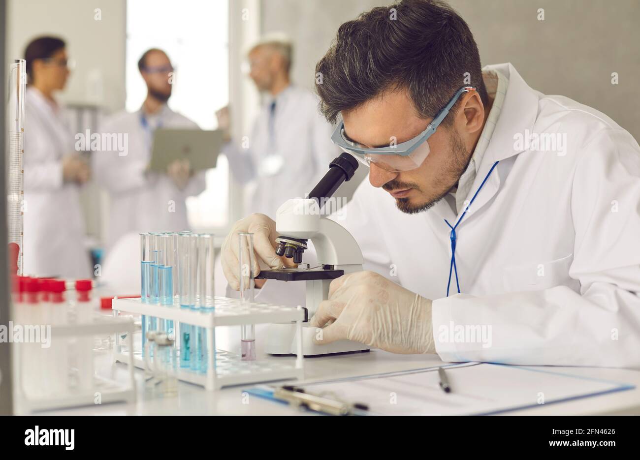 Scientifique sérieux en blouse de laboratoire, lunettes et gants regardant l'échantillon sous microscope Banque D'Images