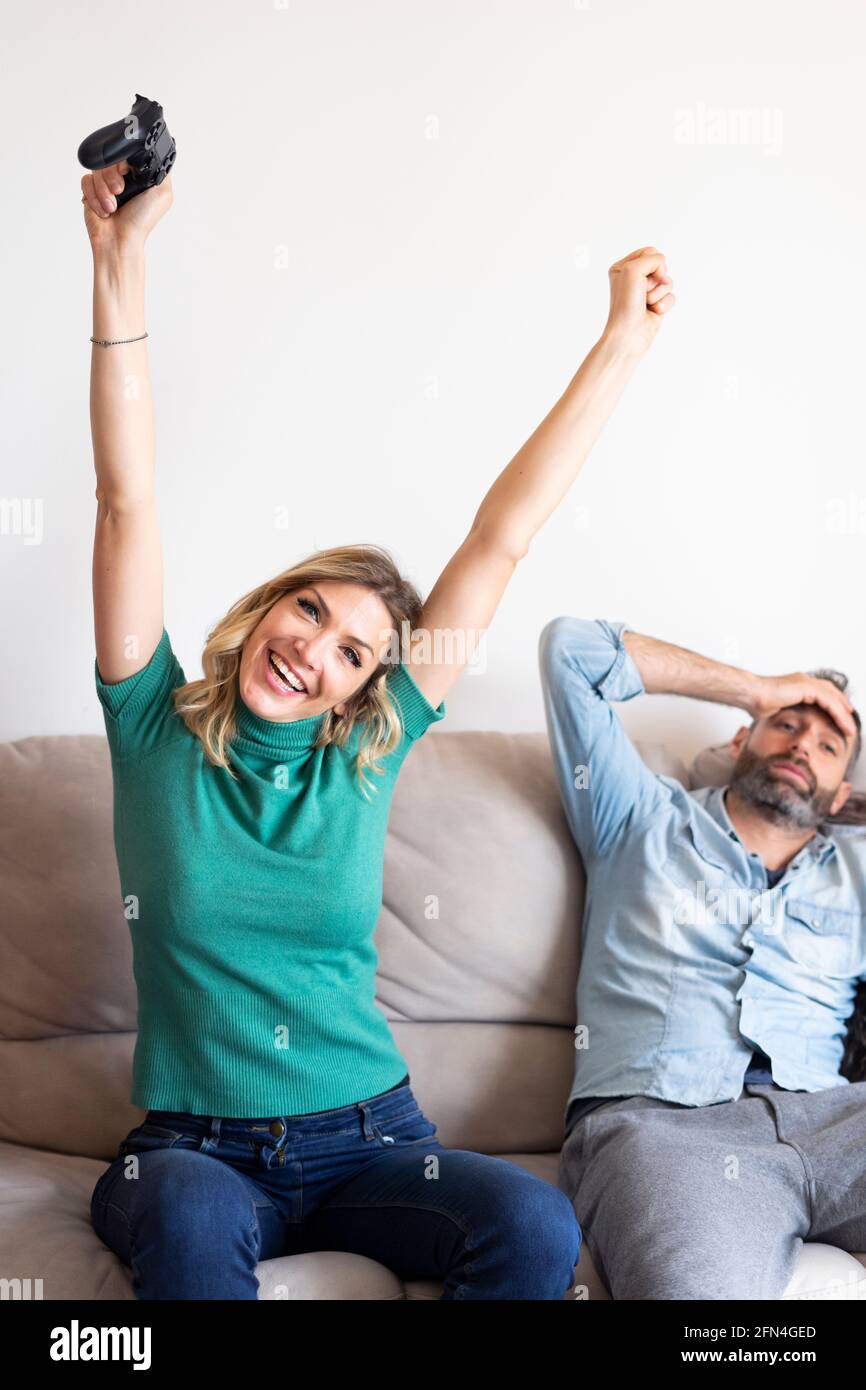 Le couple heureux joue à la console avec des manettes. La femme blonde gagne le jeu et se réjouit. Triste et fatigué mari s'effondre sur le canapé. Banque D'Images