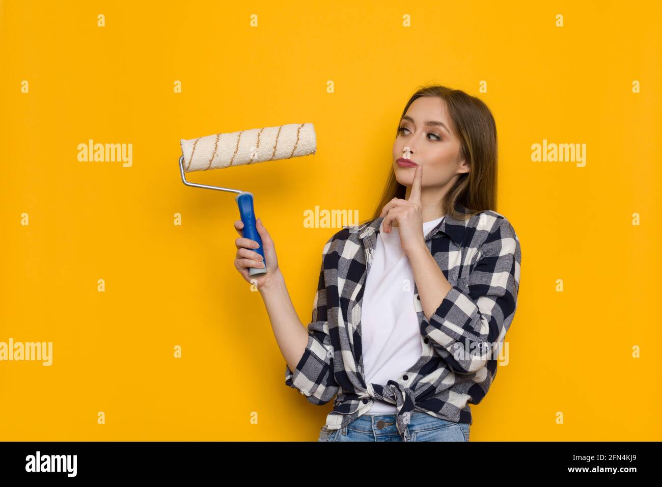 Jeune femme blonde en chemise de bûcherons tient le rouleau de peinture et de penser. Taille relevée sur fond jaune Banque D'Images