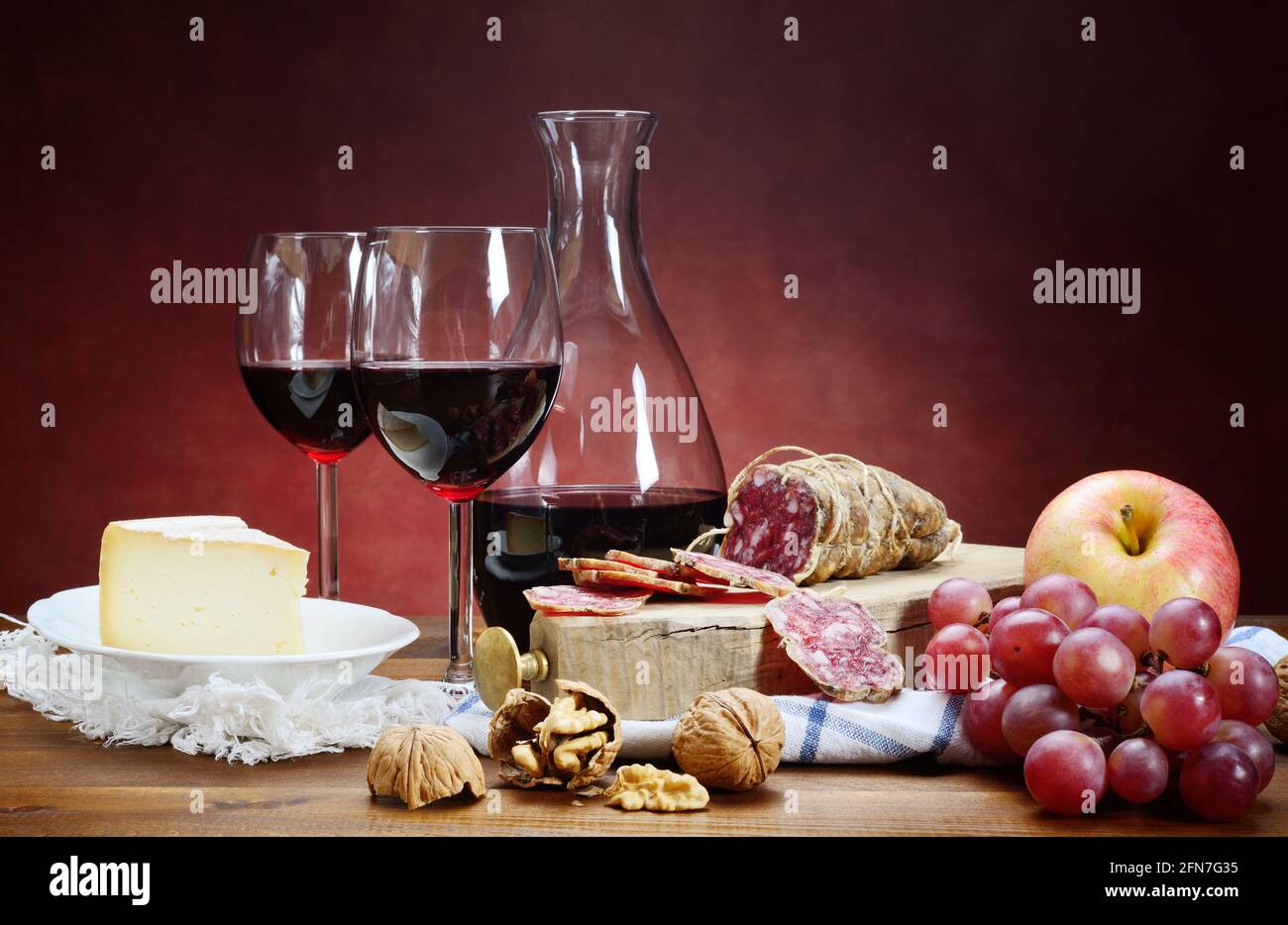 Salami, fromage, noix, raisins et vin rouge. Deux verres avec carafe sur table en bois et arrière-plan rouge avec espace pour le texte. Gros plan, vie fixe. Banque D'Images