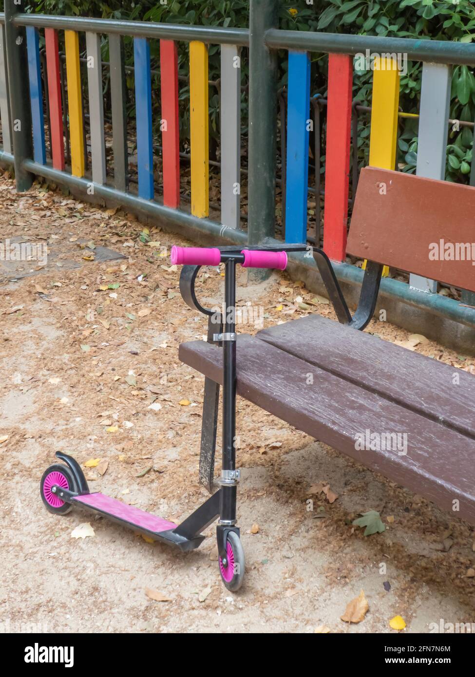 Un scooter rose pour enfants se penche sur un banc de jeu à côté une clôture multicolore Banque D'Images