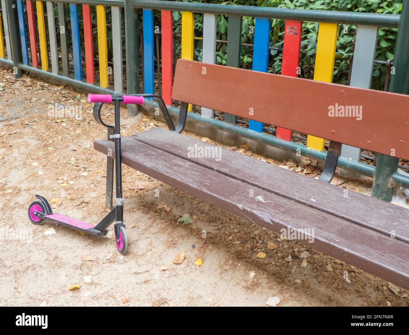 Un scooter rose pour enfants se penche sur un banc de jeu à côté une clôture multicolore Banque D'Images