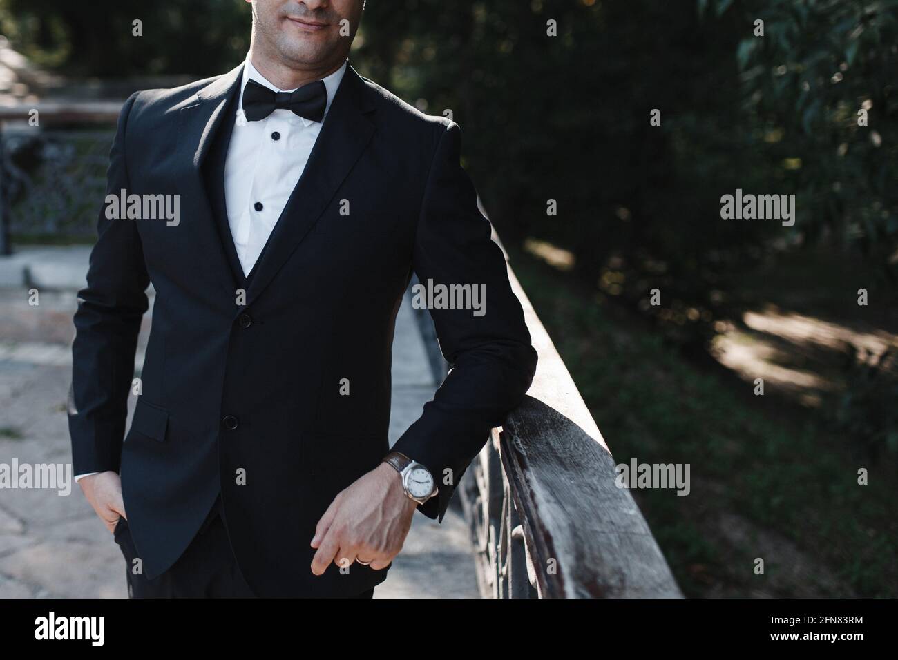 Un homme dans un costume noir classique avec un noeud papillon et une montre sur son bras s'appuie sur une main courante contre un fond d'arbres. Place pour votre texte Banque D'Images