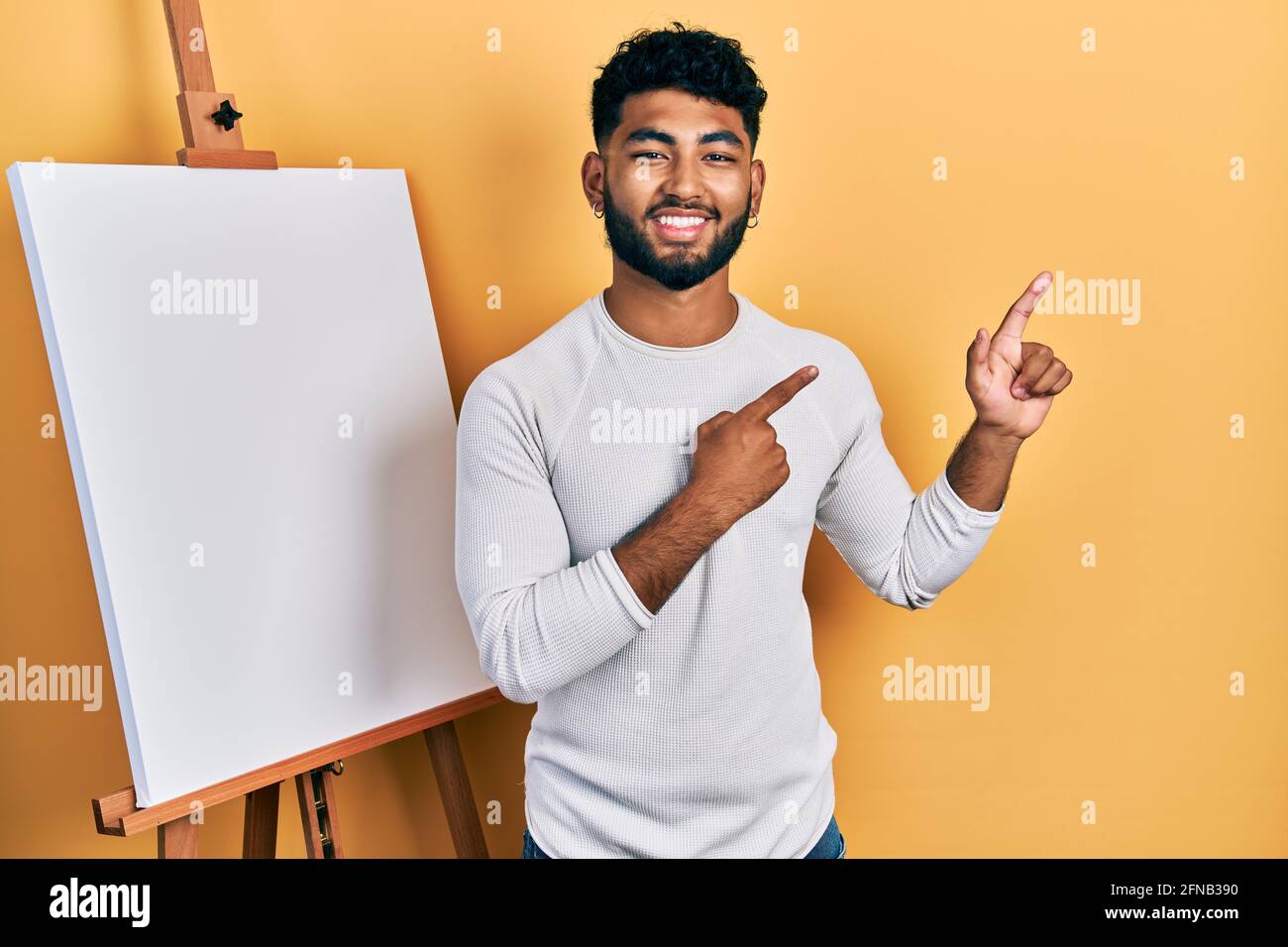 Homme arabe avec barbe debout par le chevalet de peintre stand souriant et  regardant la caméra pointant avec deux mains et les doigts sur le côté  Photo Stock - Alamy