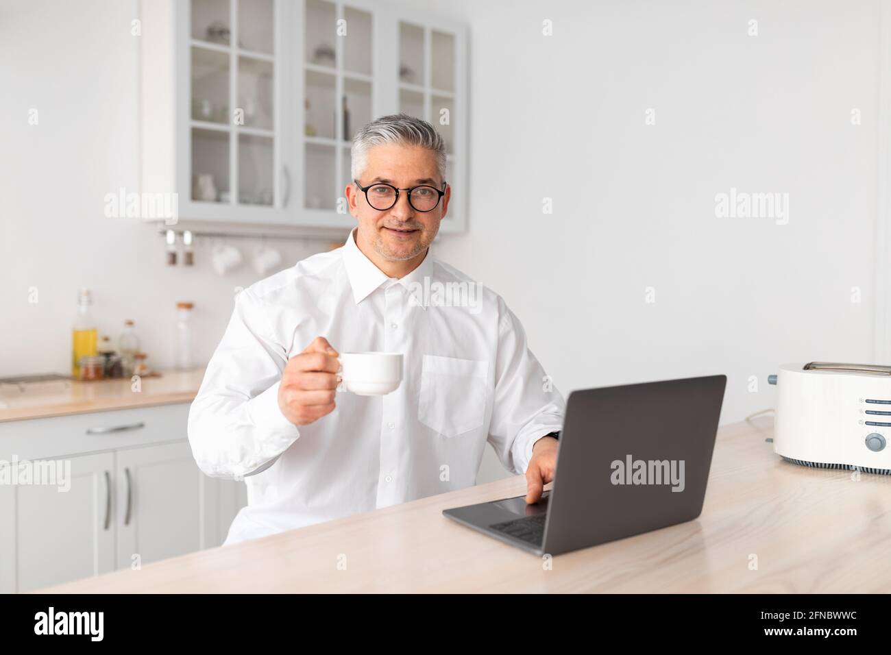 Il est temps de prendre le café du matin. Homme d'affaires mature travaillant sur un ordinateur portable et buvant une boisson chaude, assis dans la cuisine Banque D'Images