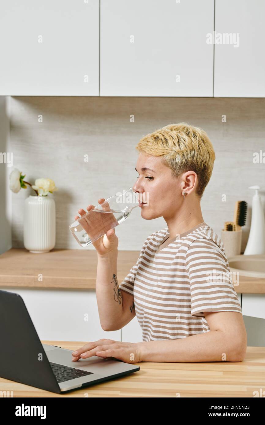 Une jeune femme blonde qui boit du verre tout en étant assise table dans la cuisine Banque D'Images