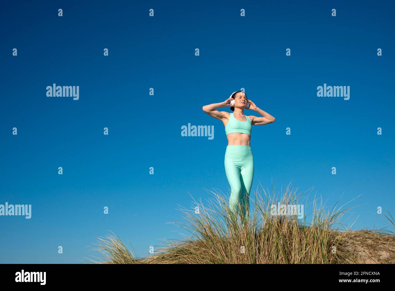 belle femme portant des vêtements de sport écouter de la musique porter des écouteurs tout en faisant de l'exercice à l'extérieur sur fond bleu ciel. Banque D'Images