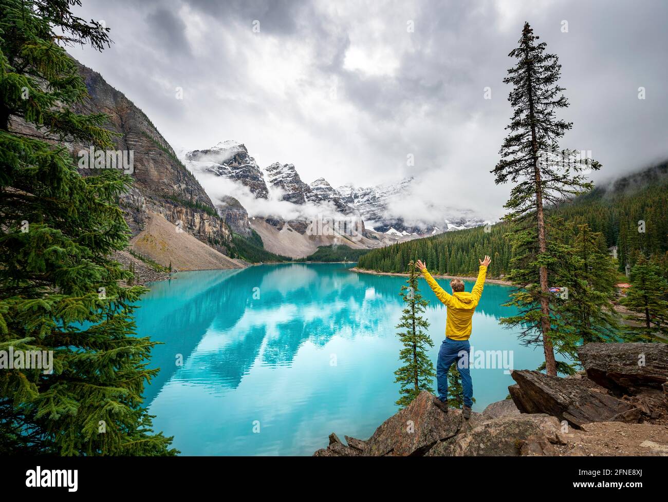 Jeune homme qui étire ses bras dans l'air, sommets de montagne nuageux, reflet dans le lac glaciaire turquoise, lac Moraine, vallée des dix sommets, Rocky Banque D'Images