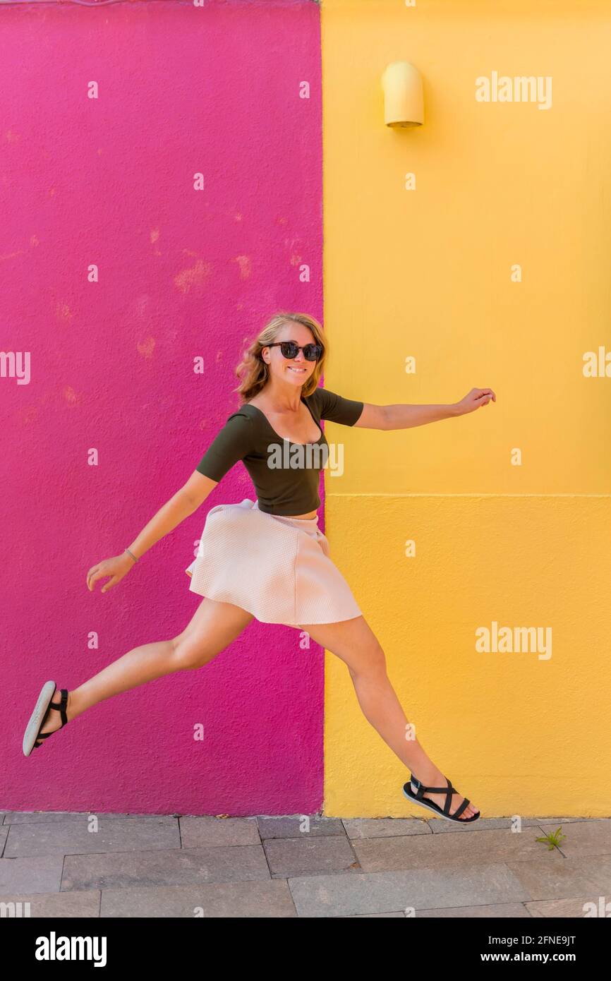Jeune femme en robe saute heureusement devant la maison colorée, façade de maison jaune et rose, île de Burano, Venise, Vénétie, Italie Banque D'Images