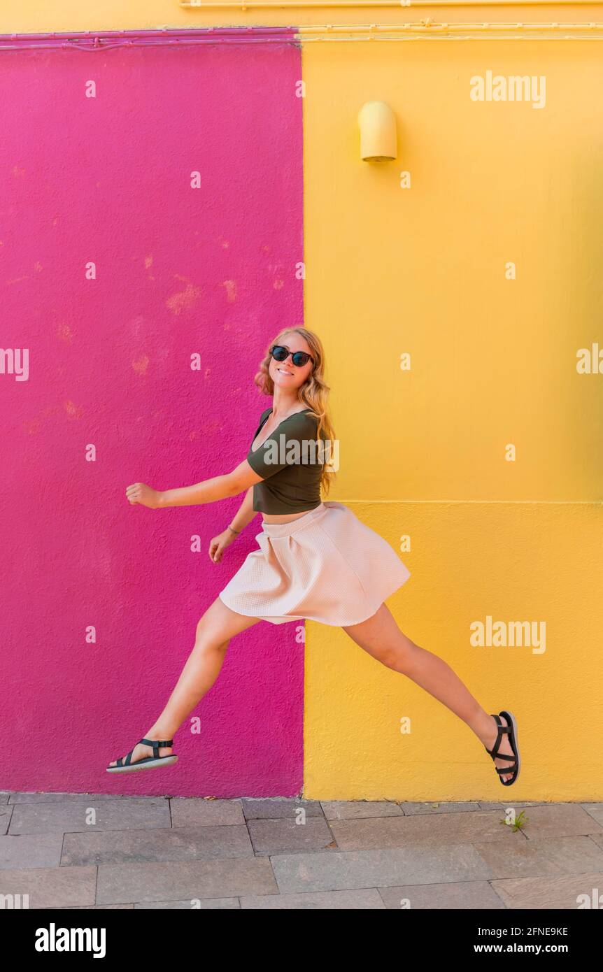 Jeune femme en robe saute heureusement devant la maison colorée, façade de maison jaune et rose, île de Burano, Venise, Vénétie, Italie Banque D'Images