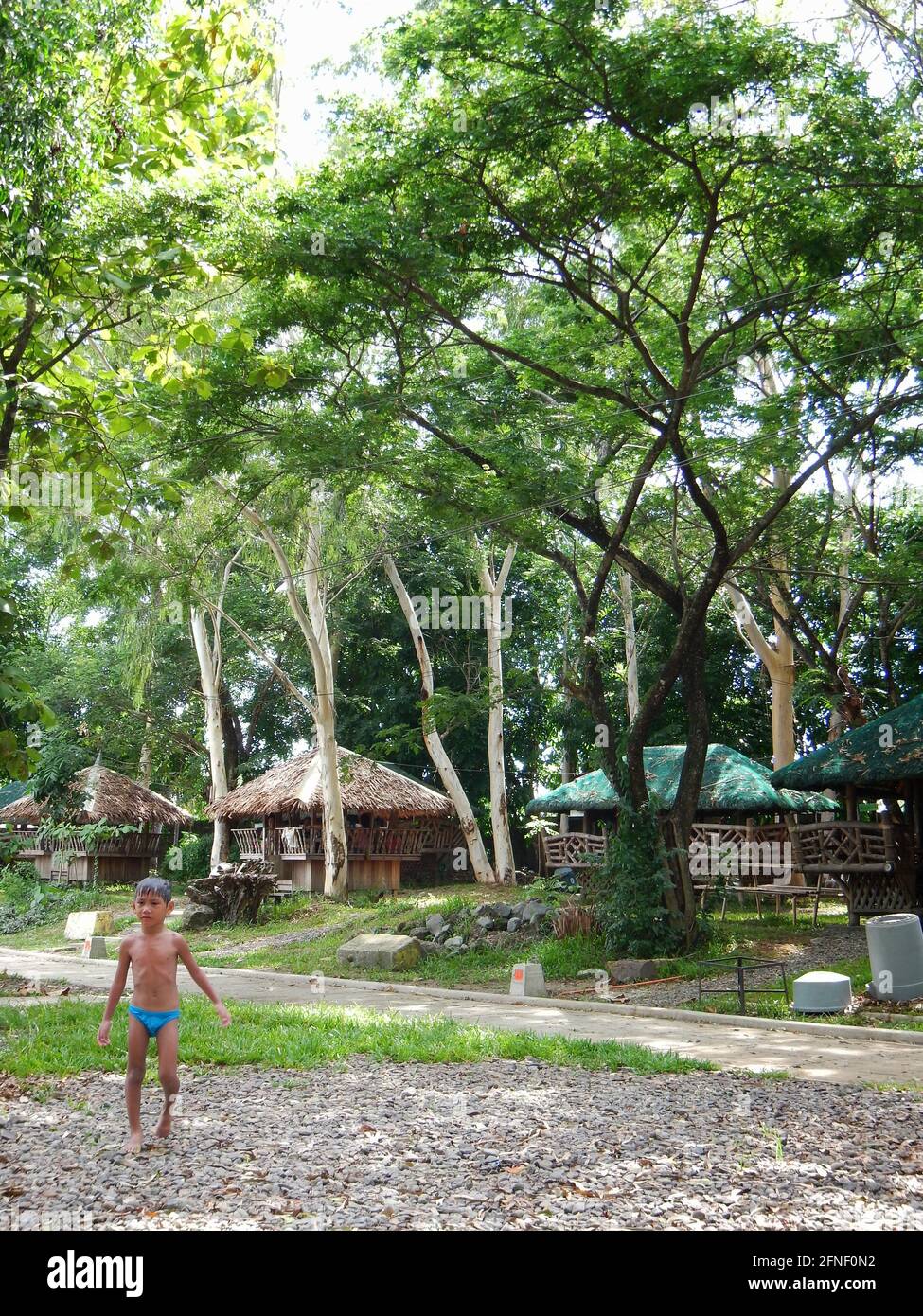 Petit garçon portant un maillot de bain bleu debout sur un chemin devant des huttes au jardin aquatique, Bacolod, Philippines Banque D'Images