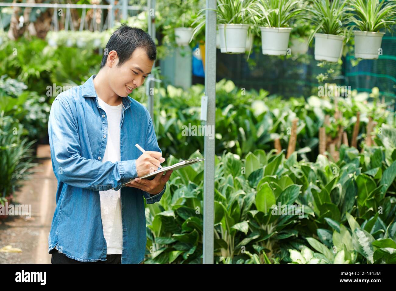 Jeune homme souriant travaillant à la pépinière, comptant les plantes et prenant des notes dans le document Banque D'Images