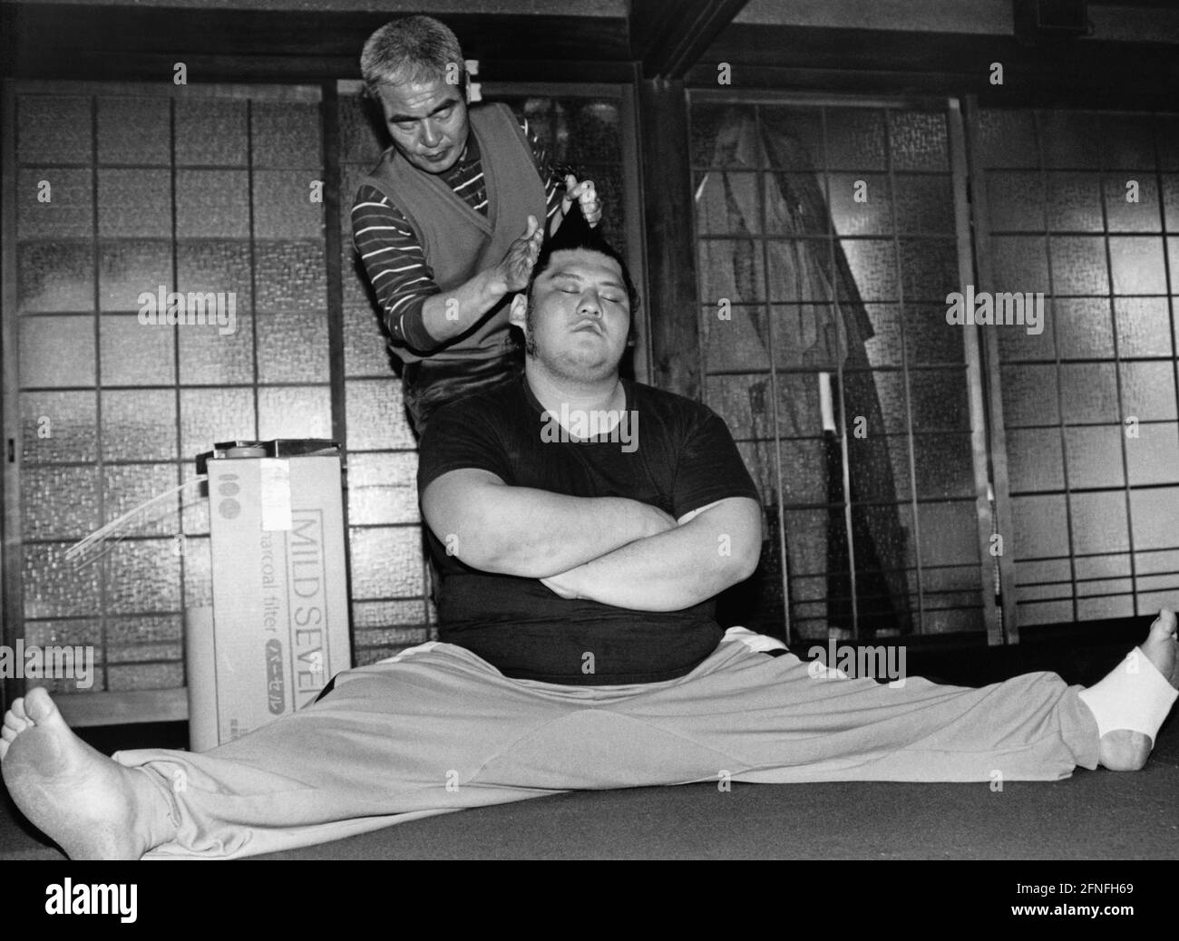 Un lutteur sumo s'assoit dans les fentes sur le sol tandis qu'un barber attache ses cheveux dans une tresse. [traduction automatique] Banque D'Images