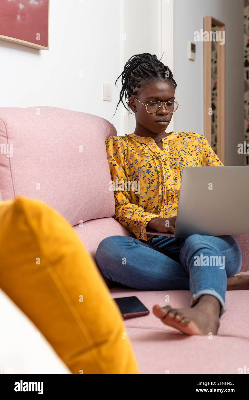 Femme indépendante afro-américaine concentrée assise sur un canapé à la maison et de taper sur netbook tout en travaillant sur le projet Banque D'Images
