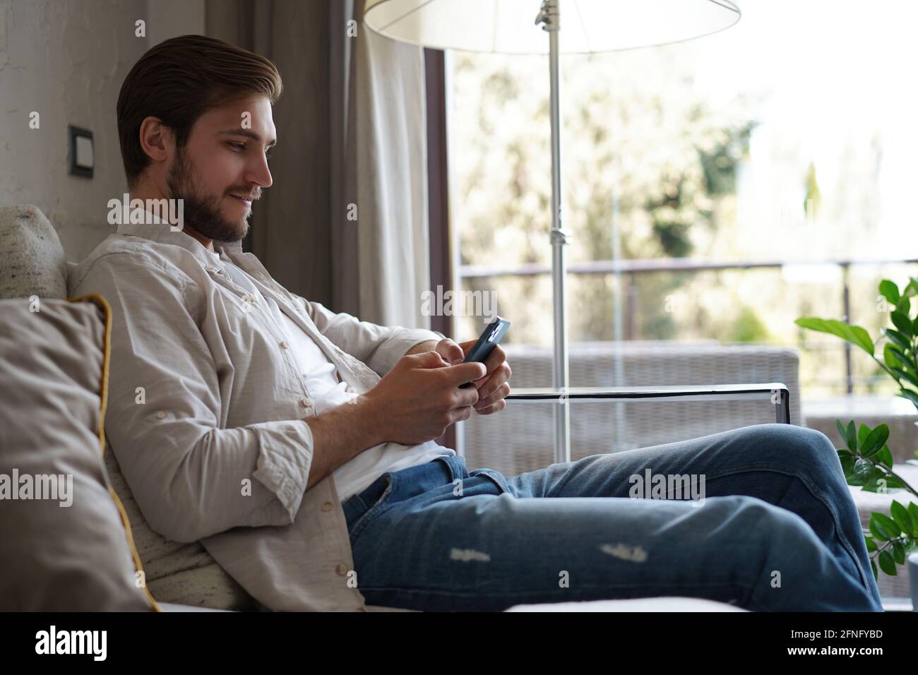 Beau homme dans un t-shirt de base souriant et tenant le téléphone portable dans les mains tout en étant assis sur un canapé dans le salon. Banque D'Images