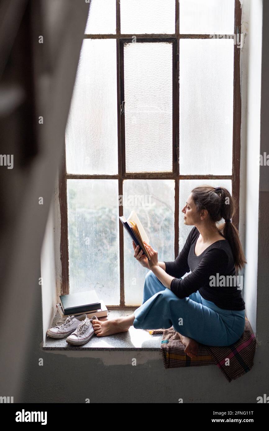 Une femme heureuse assise sur un vieux et sale rebord de fenêtre, lisant un livre. Banque D'Images