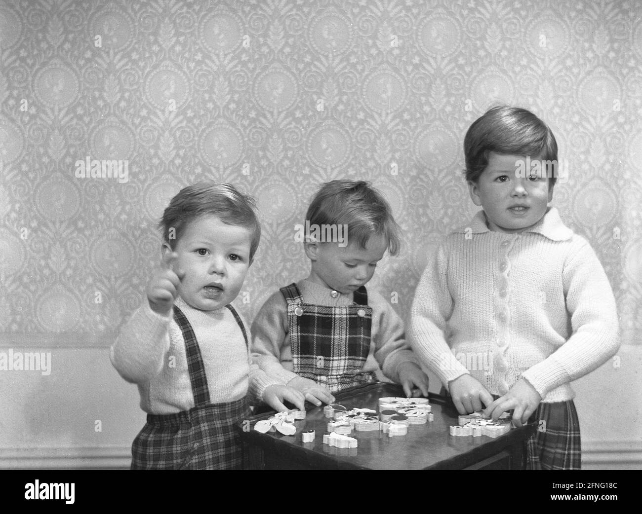 Années 1950, historique, dans leurs tenues tartan, deux petits garçons et une jeune fille, frères et sœur, debout à une table de jeu de quilles jouant avec des pièces de puzzle en bois de jouet, Angleterre, Royaume-Uni. Banque D'Images