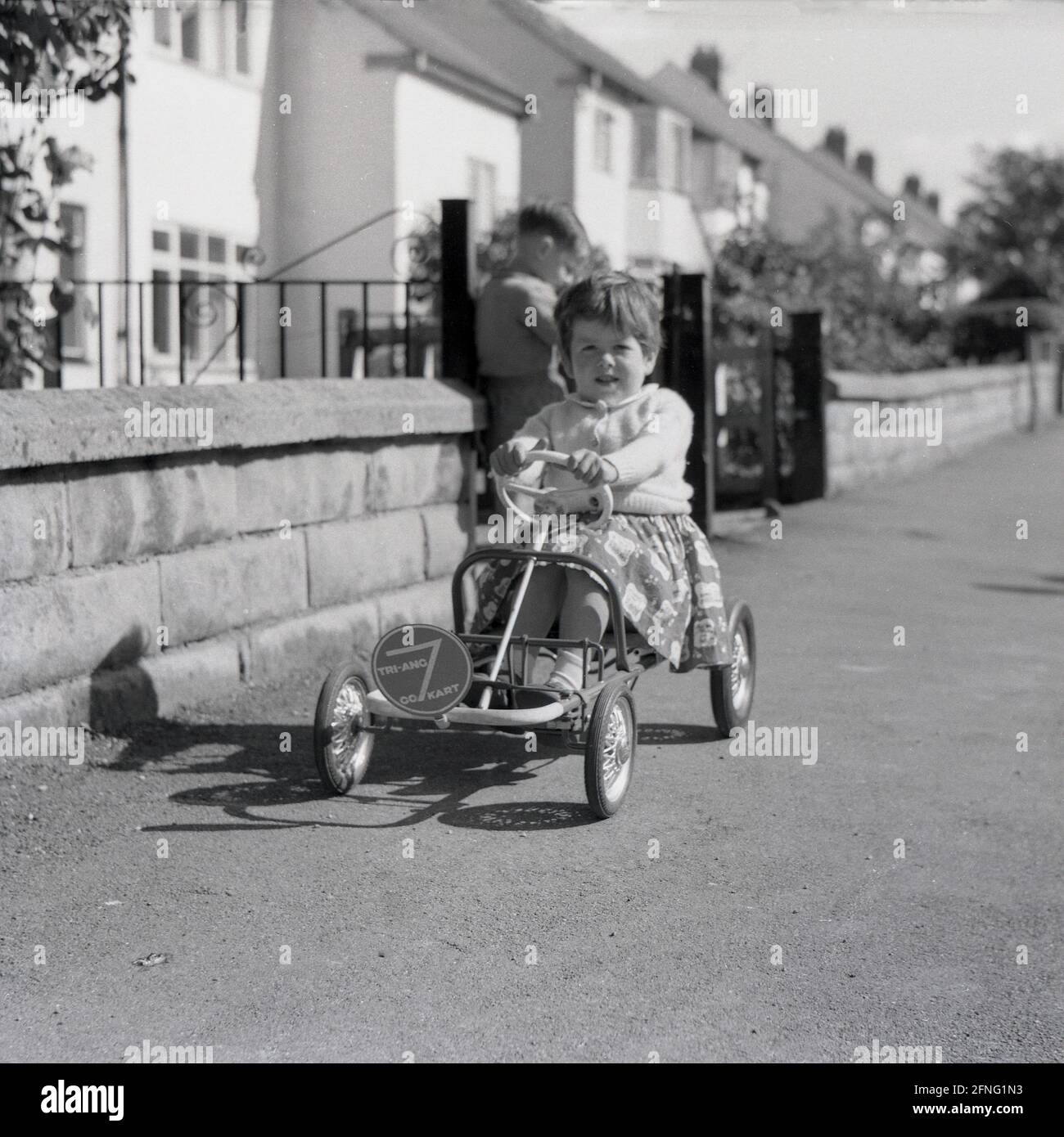 1959, historique, dehors dans la rue sur un trottoir, une jeune fille assise sur une voiture jouet à pédales à cadre métallique, un Tri-Ang 7 Go Kart, Angleterre, Royaume-Uni. Commencé en 1919, les jouets Tri-ang était le nom de marque de Lines Brothers, compan de jouets britanniques, qui étaient à un moment donné vers la fin des années 1950, considéré comme la plus grande entreprise de jouets dans le monde. Ils ont fermé en 1971. Banque D'Images