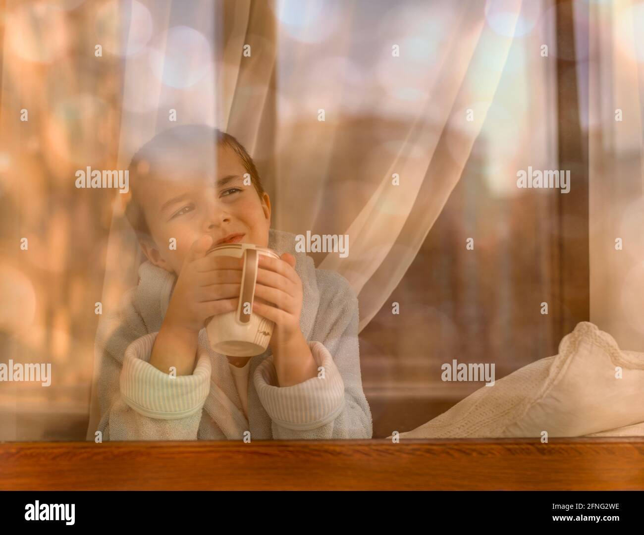 Un enfant avec une tasse dans ses mains à la fenêtre rêve de regarder la distance. Banque D'Images