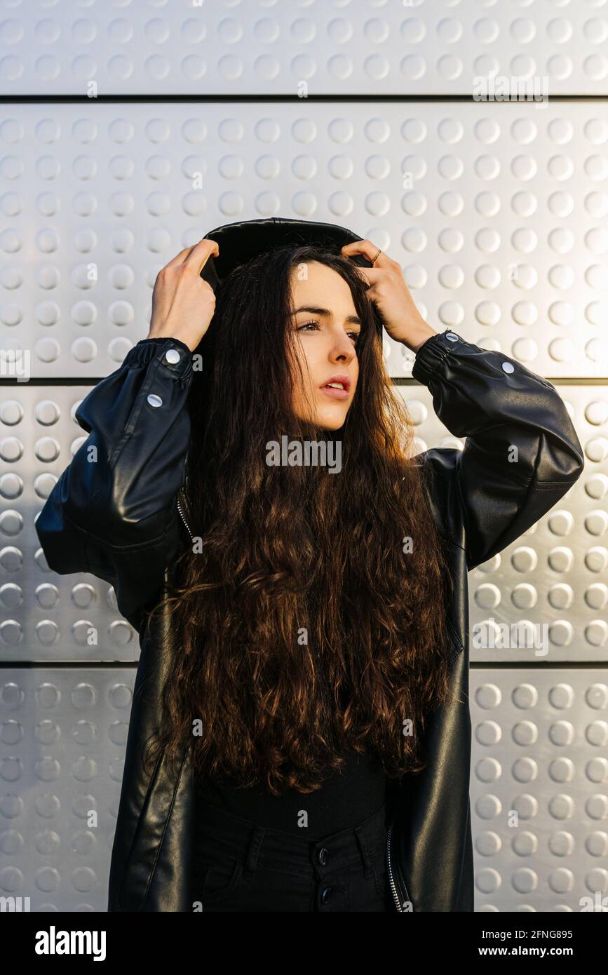 Modish jeune femme en cuir noir veste de mise sur la capuche et en  regardant loin tout en se tenant contre le mur lumineux Photo Stock - Alamy