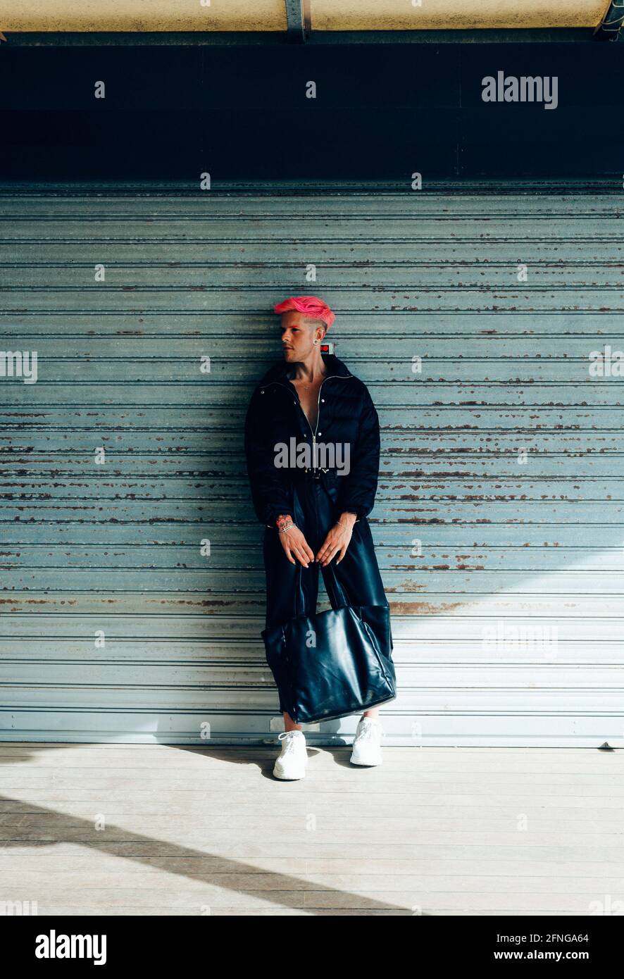 Homme transsexuel en tenue tendance avec sac à main et cheveux roses en regardant loin contre le mur abîmé par temps ensoleillé Banque D'Images