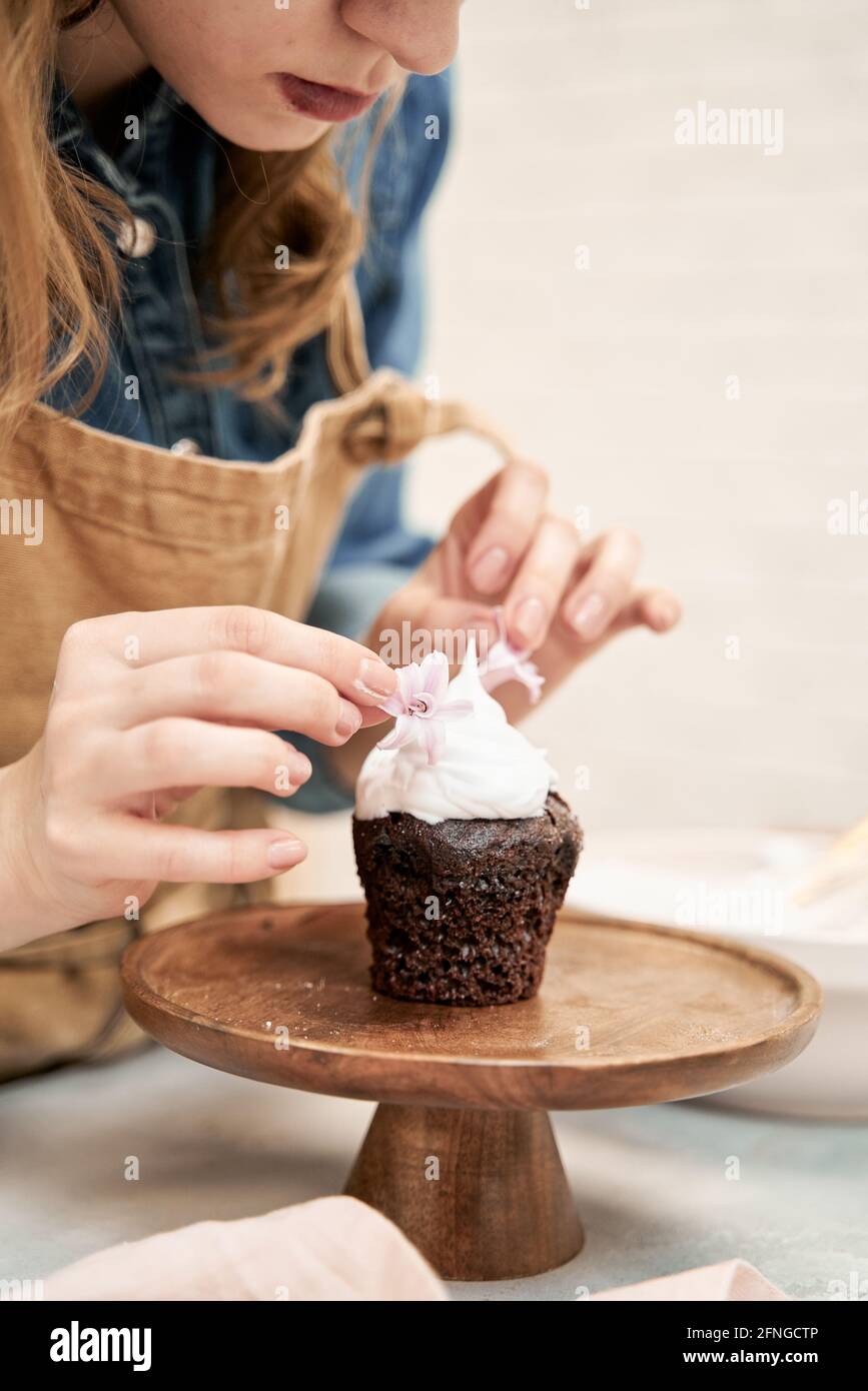 Rognez la jeune femme décorant le gâteau au chocolat avec de la crème fouettée douce et des fleurs sur un présentoir à gâteaux tout en cuisinant à la maison Banque D'Images
