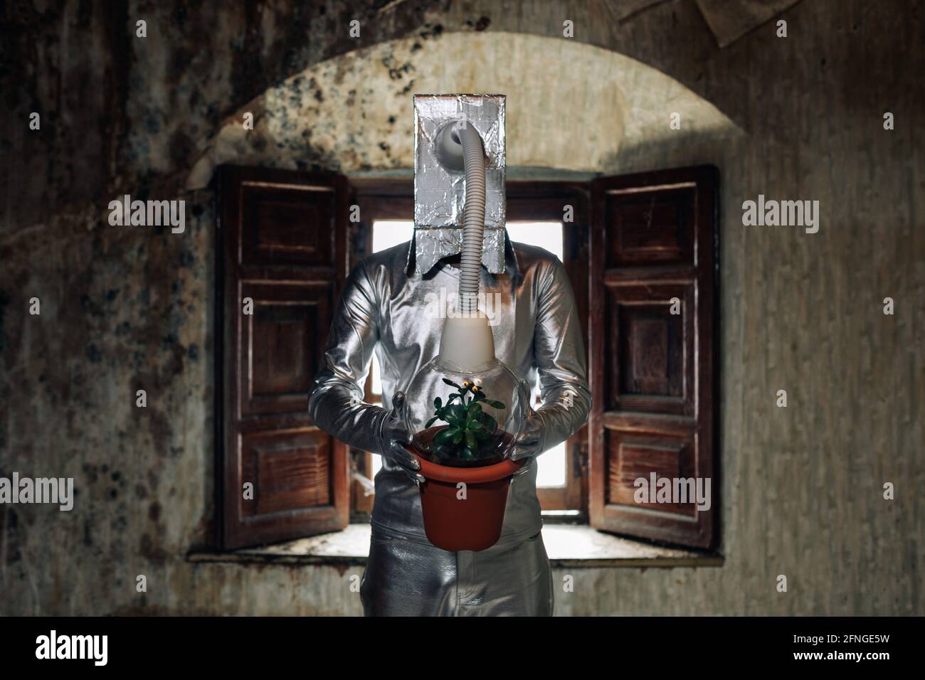 Personne non reconnaissable portant un costume argenté avec appareil respiratoire et tuyau attaché à une plante en pot debout dans une pièce abandonnée en merde Banque D'Images