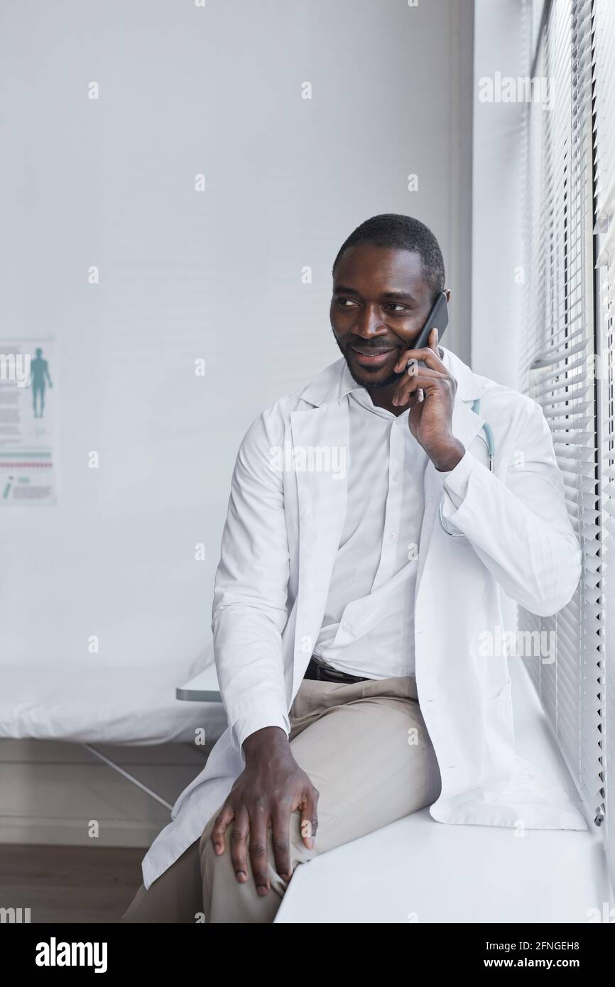 Jeune médecin africain assis près de la fenêtre et parlant dessus téléphone portable à l'hôpital Banque D'Images