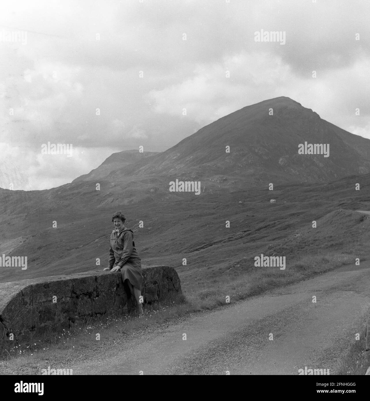 1958, historique, une dame marchée assise sur un bas mur de brique bidge sur une étroite route pavée dans les Highlands de l'Écosse. Banque D'Images